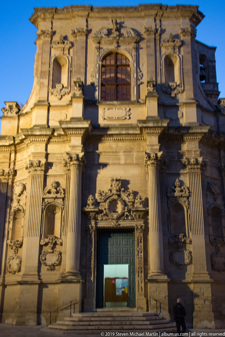 Illuminated Baroque church façade with paired columns, carved portal, niches, heraldic reliefs, and a large arched upper window.