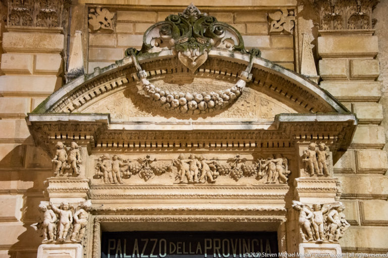 Richly carved stone pediment, garlands, shield, and figurative frieze above the Palazzo della Provincia entrance.