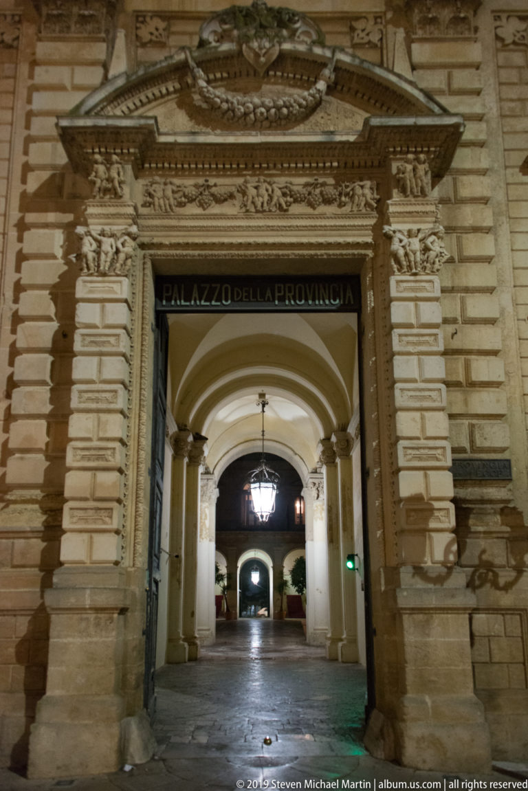 Ornate stone entrance labeled PALAZZO DELLA PROVINCIA opening into a lantern-lit vaulted passage.
