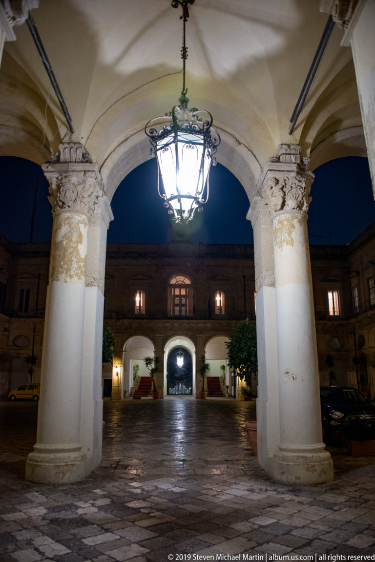 Palazzo della Provincia courtyard seen between two large columns, with a hanging lantern and illuminated arches beyond.
