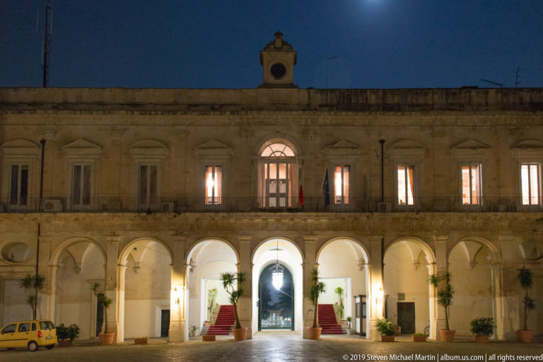 Symmetrical palazzo courtyard façade with illuminated arches, upper windows, roofline clock, and central passage at night.