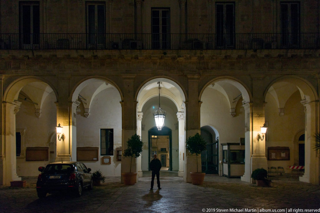 Arcaded palazzo courtyard at night with central hanging lantern, potted trees, parked car, and one standing person.