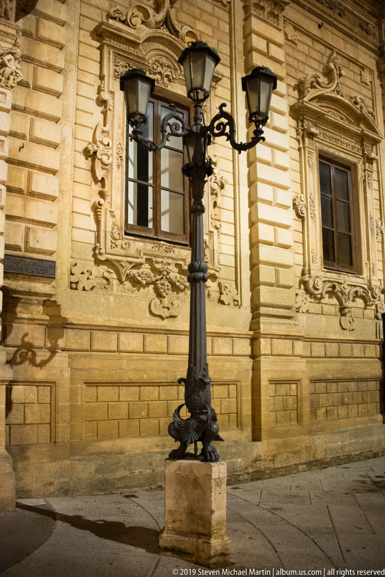 Ornate black multi-lantern streetlamp in front of carved limestone windows and rusticated palazzo wall at night.