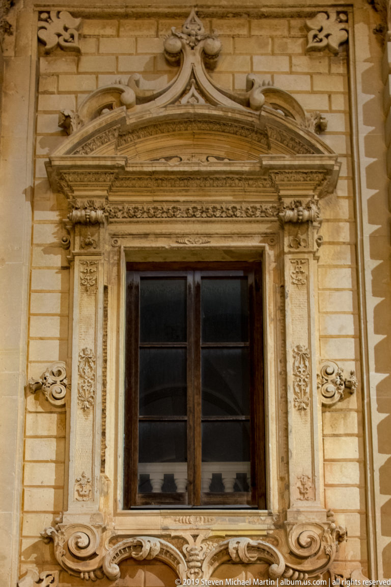 Dark wooden window framed by richly carved limestone scrolls, floral ornament, and a broken curved pediment.