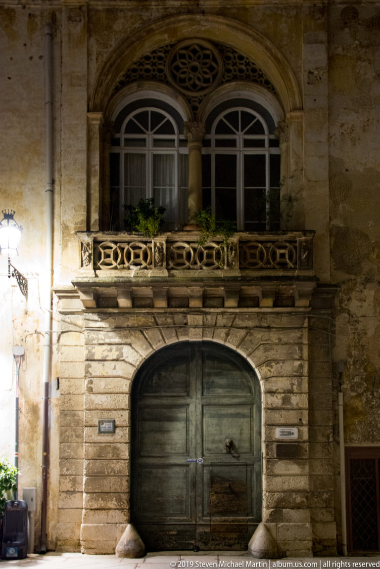 Large arched wooden doorway beneath an ornate stone balcony with paired windows and circular tracery at night.