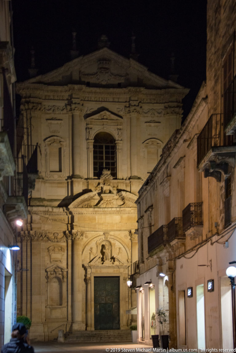 Illuminated Baroque church façade at the end of a narrow street lined with shops and balconies.