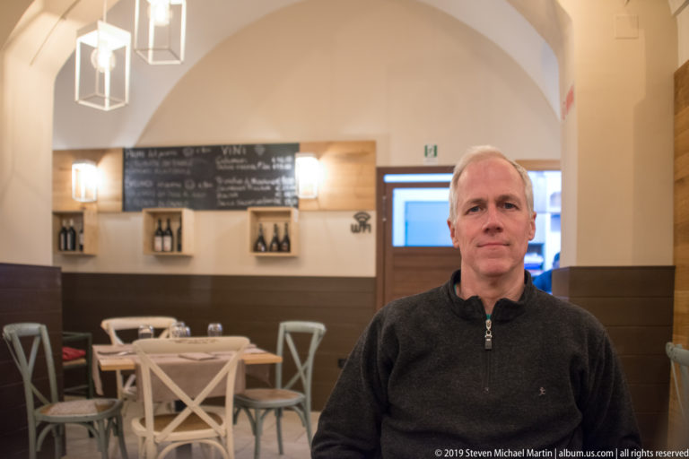 Man seated facing the camera in a vaulted restaurant dining room with white chairs and a chalkboard menu behind him.