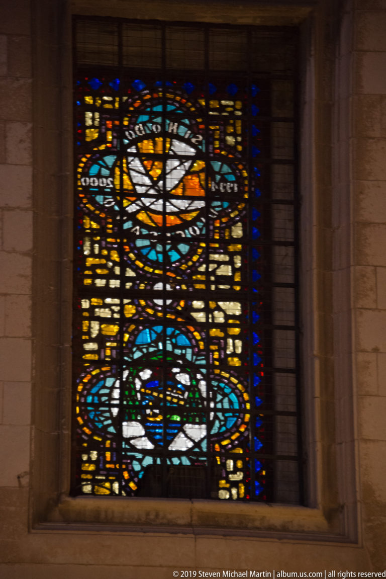 Stained-glass window in a deep stone reveal with a white dove above abstract blue, amber, red, and green forms.