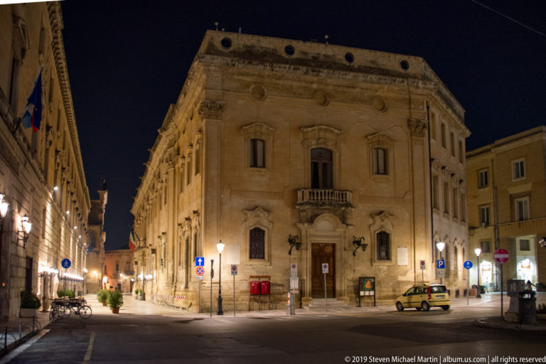 Large illuminated stone palazzo with carved windows and central balcony at a quiet Lecce street intersection.