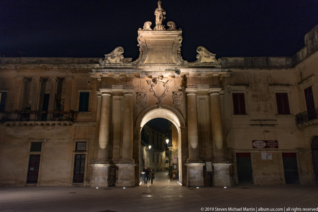 Illuminated Porta Napoli with paired columns and a single arched passage opening onto a narrow street at night.