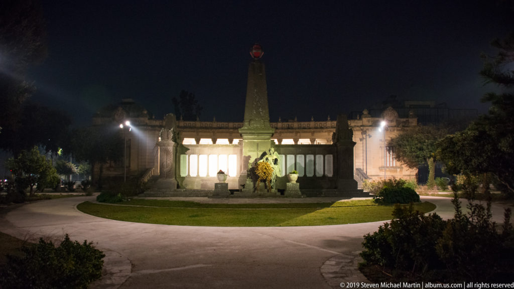 Frontal nighttime view of a tall stone monument, sculpted pedestal, curved paths, lawn, and illuminated colonnade.