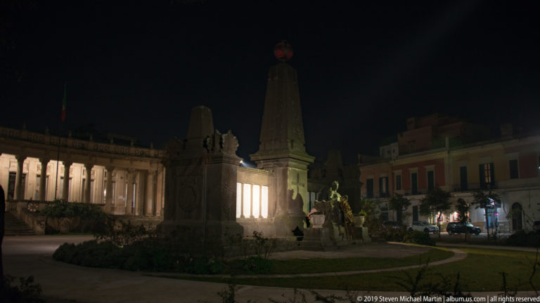 Night view of a tall stone monument with sculpted base between a curved colonnade and nearby city buildings.