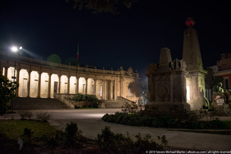 Tall stone monument with sculpted base in a landscaped space, backed by a brightly illuminated curved colonnade.
