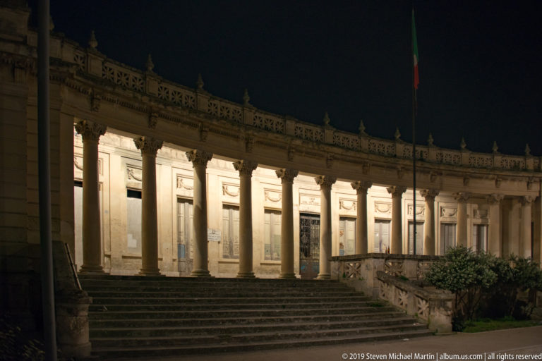 Curving illuminated colonnade behind broad steps, with repeated columns, a flagpole, and low landscaping at night.