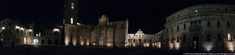 Wide nighttime streetscape with curved stone façade at right and illuminated historic buildings receding into the distance.