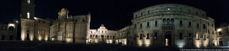 Night view of a Lecce square with a rounded stone corner building, older façades, and illuminated arcades.