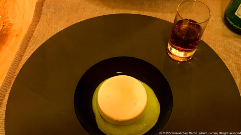 Round custard dessert in green sauce beside a small glass of amber digestif on a black restaurant table.