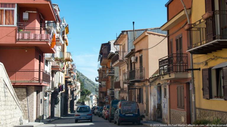 A narrow Monreale street between warm-colored buildings leads toward a distant green hillside.