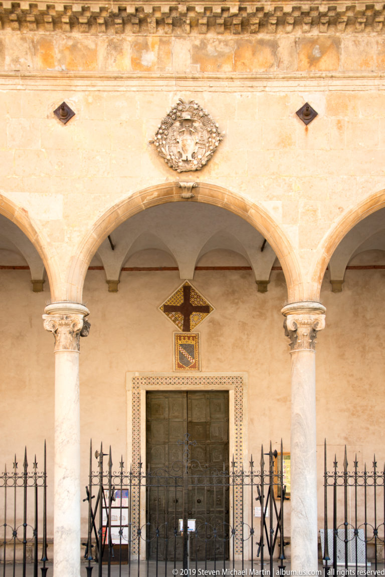 A dark doorway beneath pale arches is marked by a cross and screened by a low metal barrier.