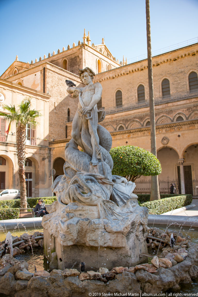 The Fontana del Tritone and its basin in Piazza Vittorio Emanuele II, framed by palms and the surrounding buildings.