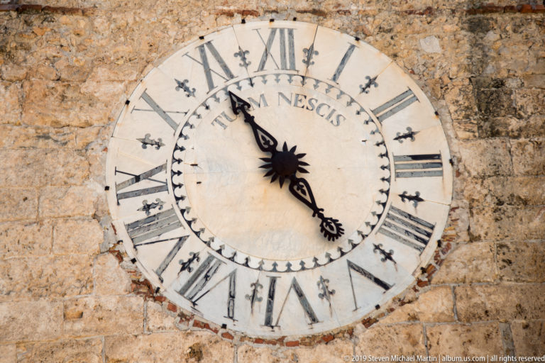 A pale circular clock with dark Roman numerals and hands is mounted on a weathered stone wall.
