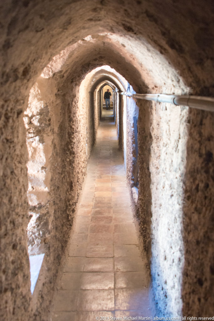 A narrow stone passage runs beneath a sequence of low arches toward a brighter opening.