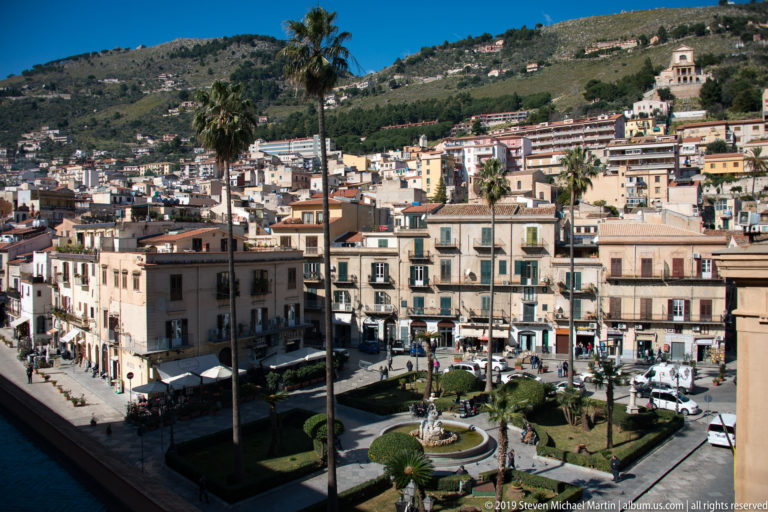 A palm rises above Monreale’s hillside neighborhood, where dense buildings climb toward a green ridge.