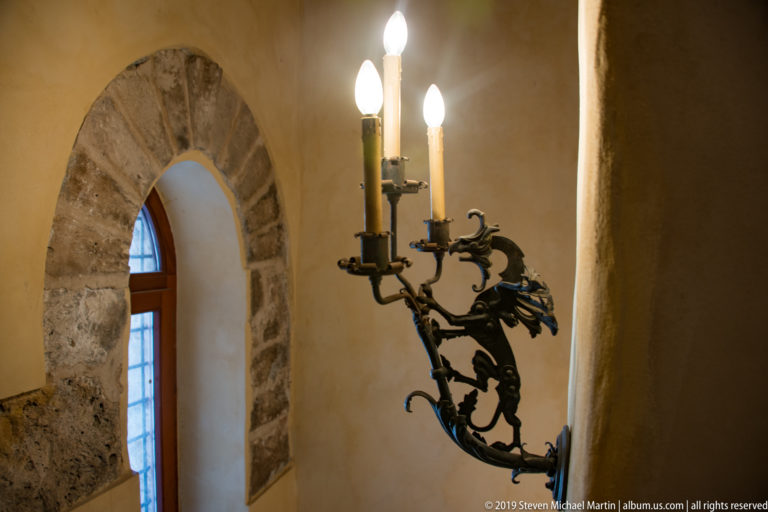 A multi-arm black metal sconce projects beside a narrow arched window in a stone wall.