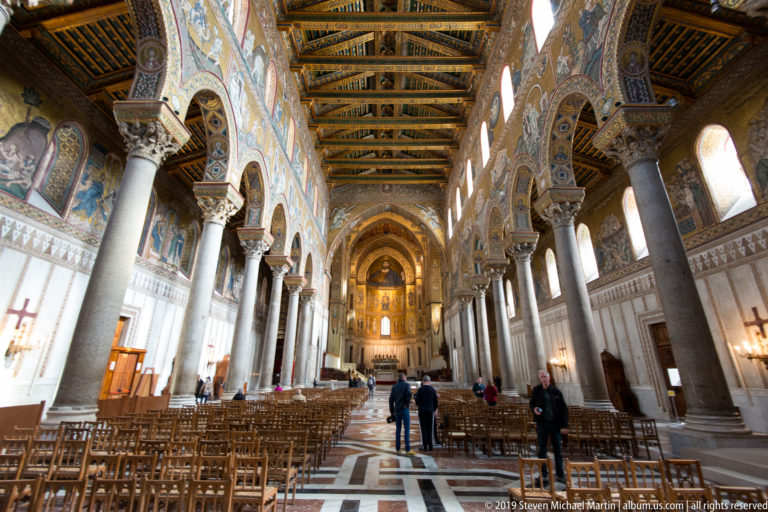 Visitors stand among rows of chairs in the cathedral nave, facing the distant mosaic sanctuary beneath tall arches.