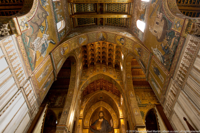 Low-angle interior view of Monreale Cathedral’s gilded apse mosaic, framed by painted arches and ceiling decoration.