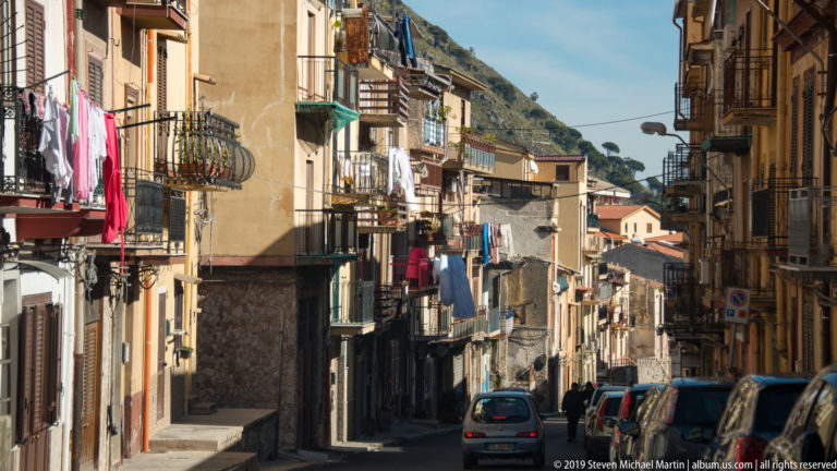 A narrow Monreale street runs between close-set apartment buildings with balconies and parked cars.