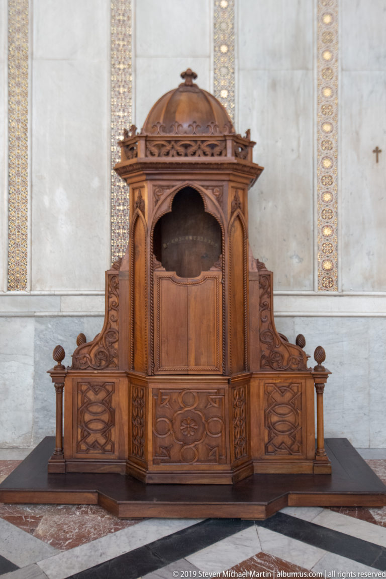 A carved wooden structure with a domed canopy and central recess stands against a pale wall.