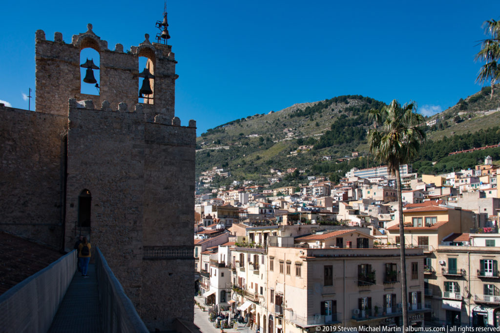 A shaded cathedral wall frames bright hillside houses, a palm, and green slopes in Monreale.
