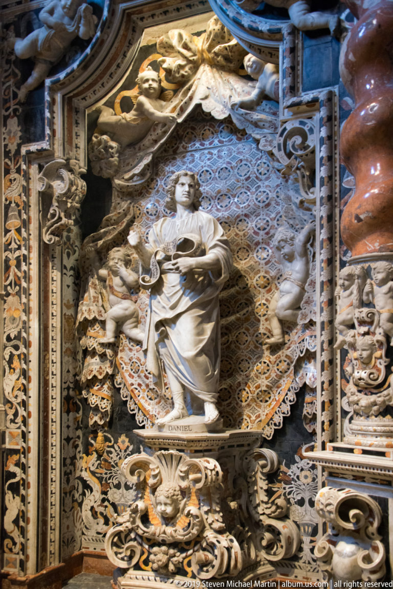 Daniel stands with a scroll in a richly patterned niche beside a smaller kneeling figure.