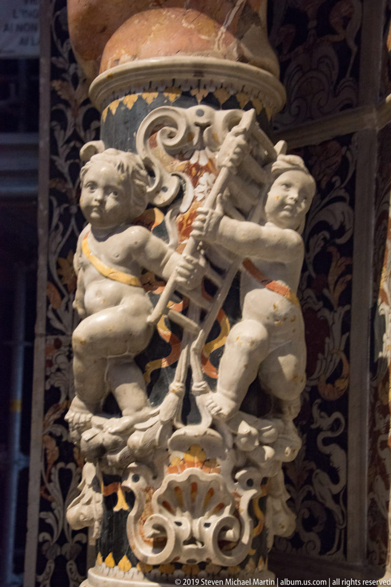 A pale winged figure stands above a carved face within a dark ornamental frame.