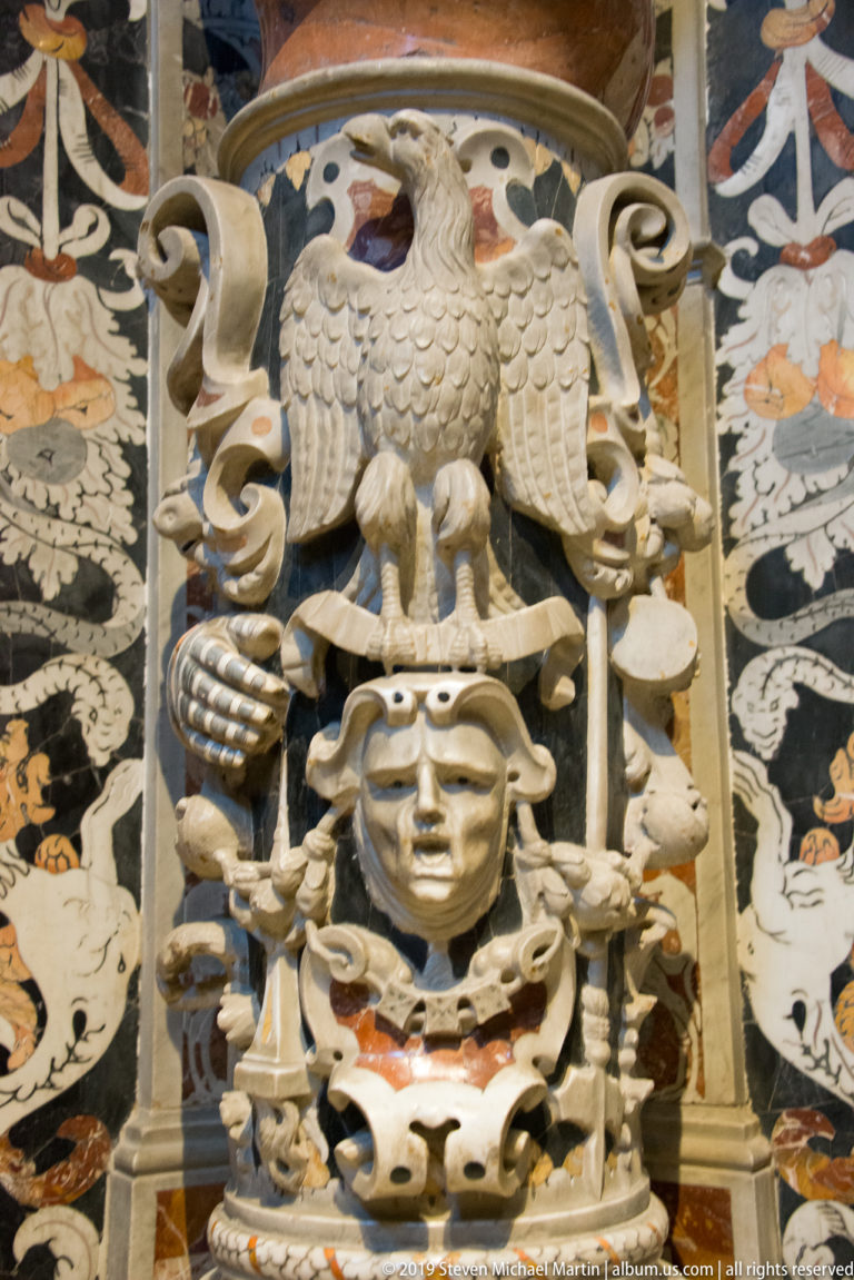A winged carved figure supports a marble prophet statue amid geometric ornament.