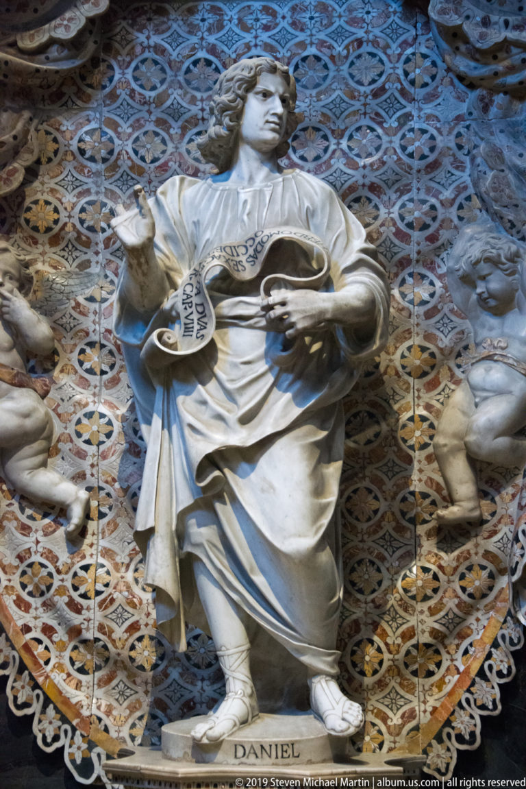 A marble figure labeled “DANIEL” holds a scroll before a blue and gold patterned niche.
