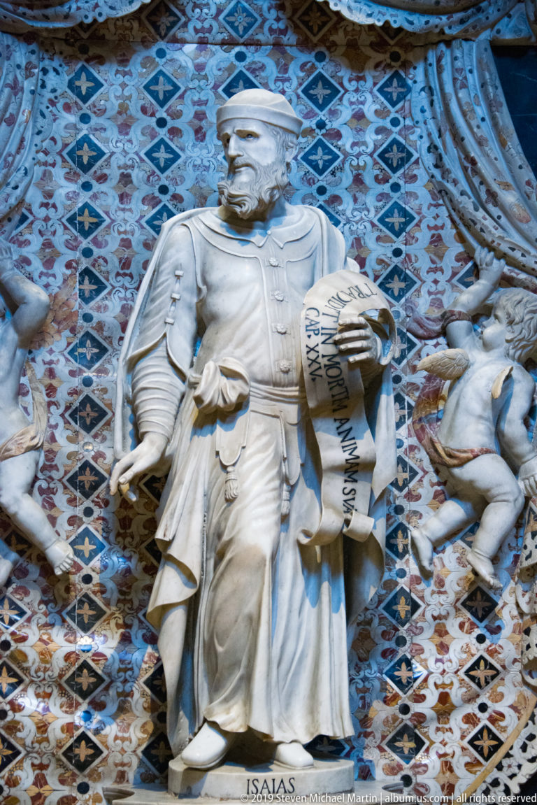 A marble prophet labeled “ISAIAS” holds a scroll before a blue geometric background.