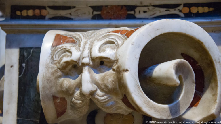 A pale carved bearded face is framed by curling stone forms and a smaller mask.