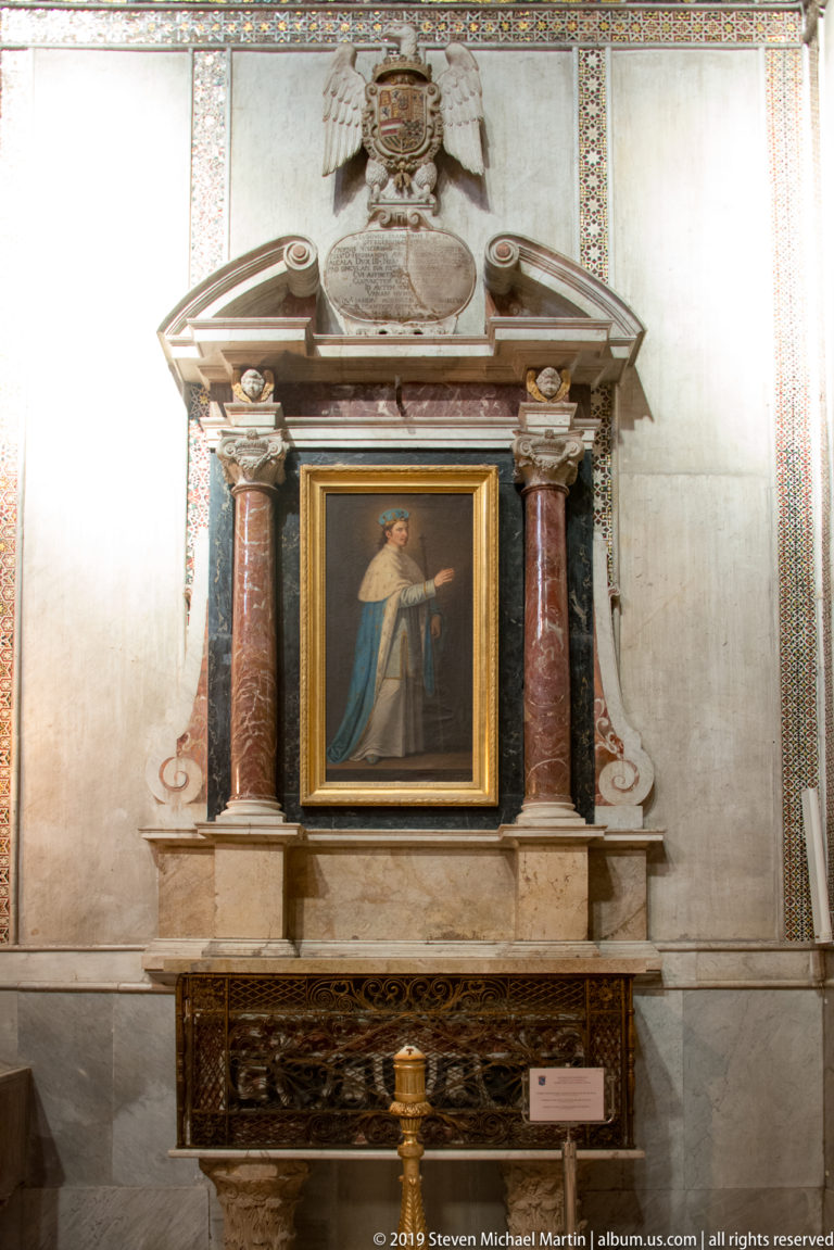 A small painting of a veiled woman hangs within a dark columned frame on a pale wall.