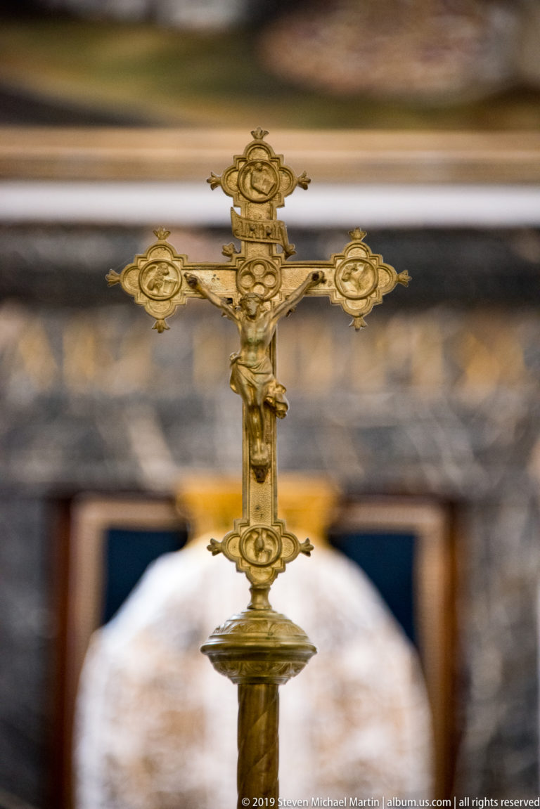 A gold-colored processional cross stands upright against a softly blurred interior.
