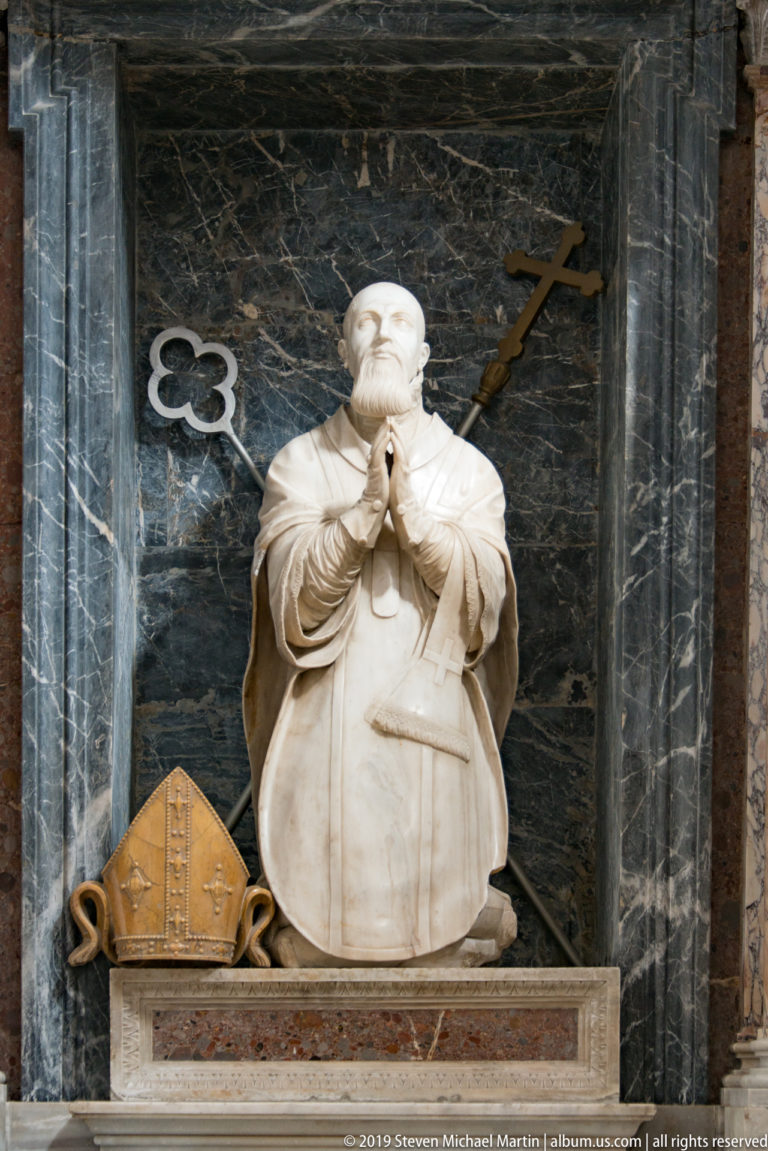 A bearded marble church figure stands in a dark niche beside a carved mitre.