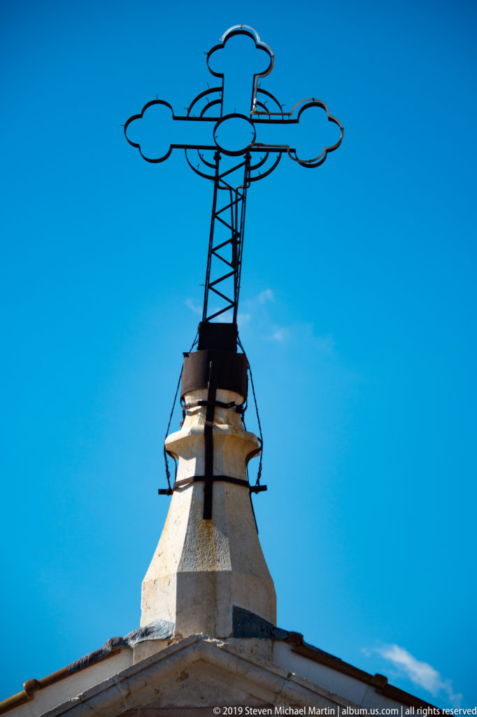 A dark ornamental metal finial rises from a pale rooftop against a clear blue sky.