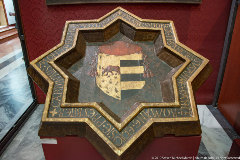 A shield-shaped emblem with a double-headed eagle and striped field is set within a blue-and-gold border.