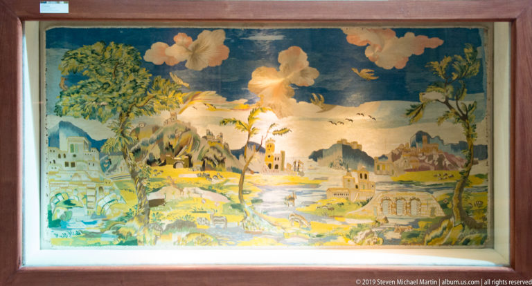 A framed mosaic landscape shows trees, buildings and water beneath a deep blue sky.