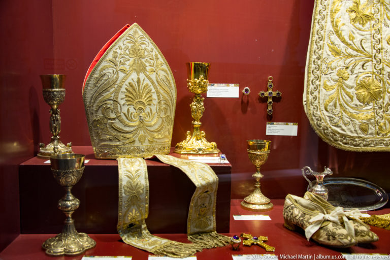 Embroidered vestments and gold-toned liturgical vessels, including a cross and chalices, are arranged in a display case.
