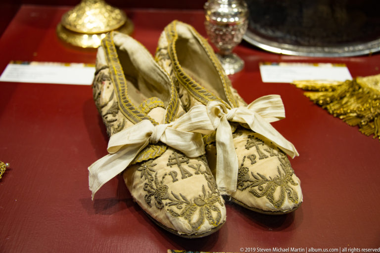 A pair of cream episcopal shoes with gold embroidery and white ribbons, displayed against red fabric.