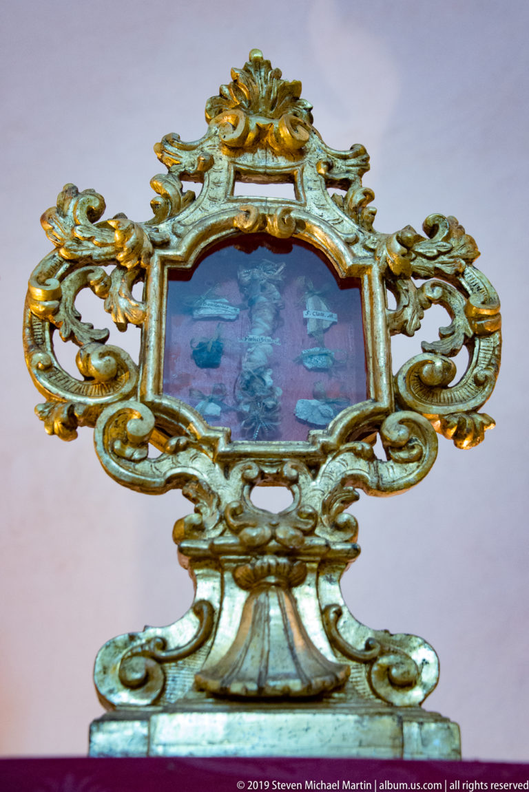 A small blue-centered devotional object is enclosed by an ornate silver-colored frame with curling ornament.