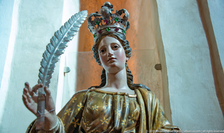 A crowned figure with dark curls and a patterned gold garment raises one hand against a pale interior background.