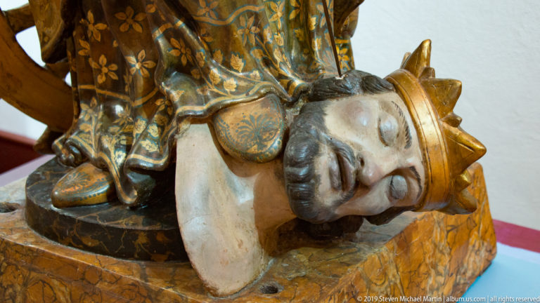 A pale sculpted face tilts downward beneath a blue and gold headdress, with a carved hand near the cheek.